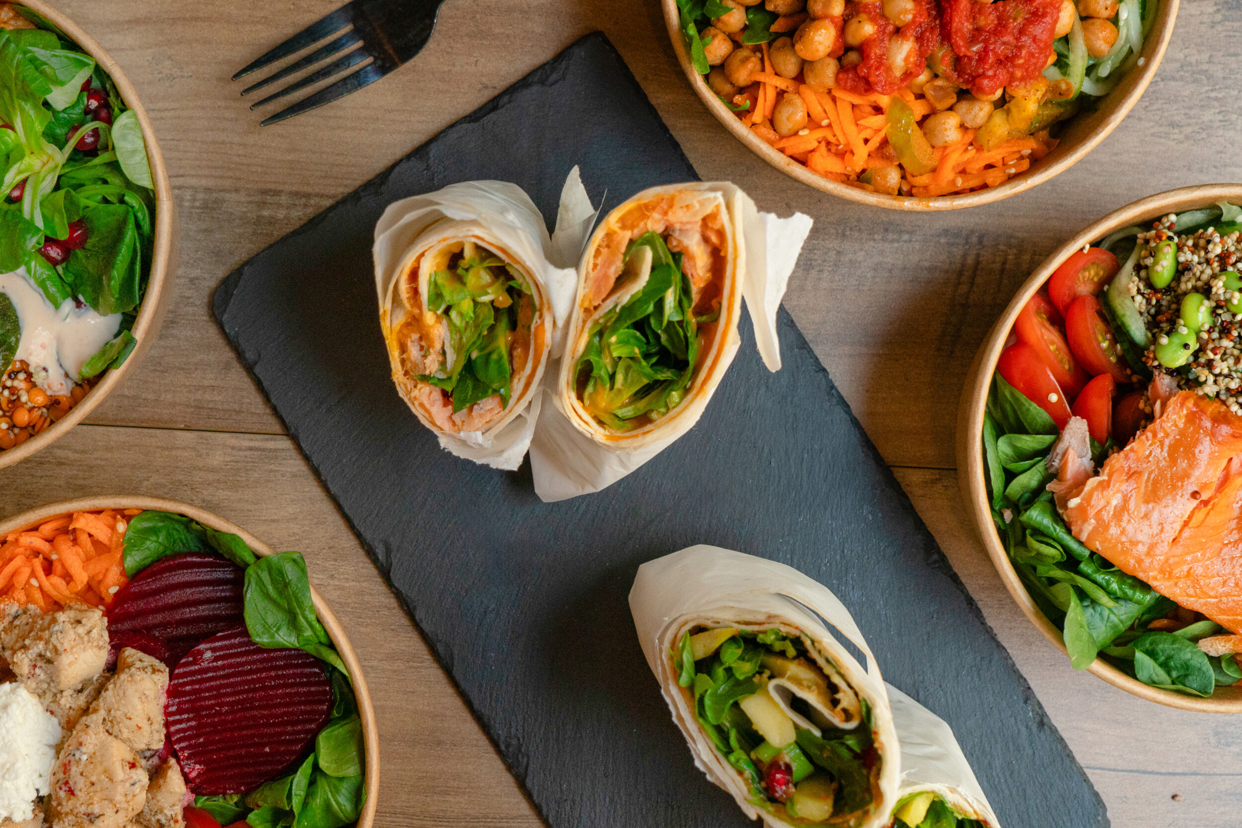 Eine bunte Auswahl an gesunden Gerichten von Mames Bowls & Wraps Gera, fotografiert als Flatlay: In der Mitte ein aufgeschnittener Lachs-Wrap, umgeben von verschiedenen Bowls mit Lachs, Hähnchen, Roter Bete und Kichererbsen auf einem Holztisch.