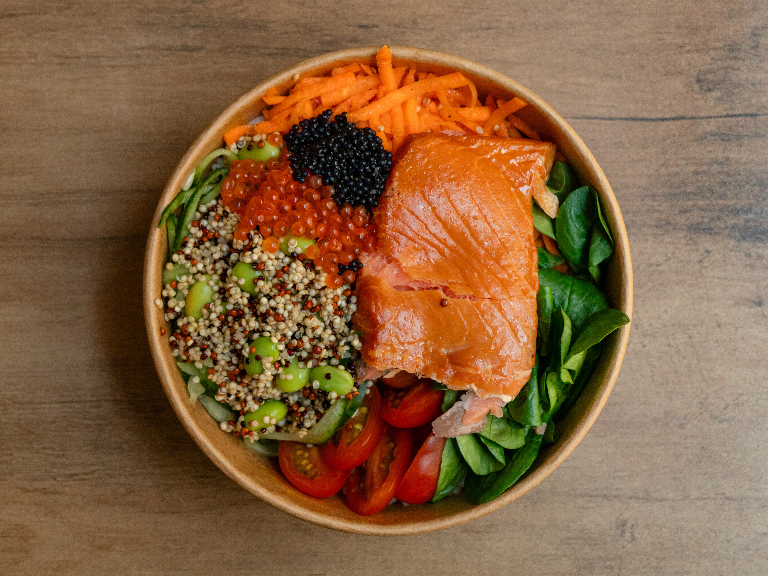 Draufsicht der Ma Kai Bowl von Mames Bowls & Wraps Gera: Eine gesunde Bowl mit einem großen Lachsfilet, Quinoa, Edamame, Karottenstreifen, Tomaten und Lachs-Kaviar, serviert in einer Holzschale. Auch glutenfrei erhältlich.