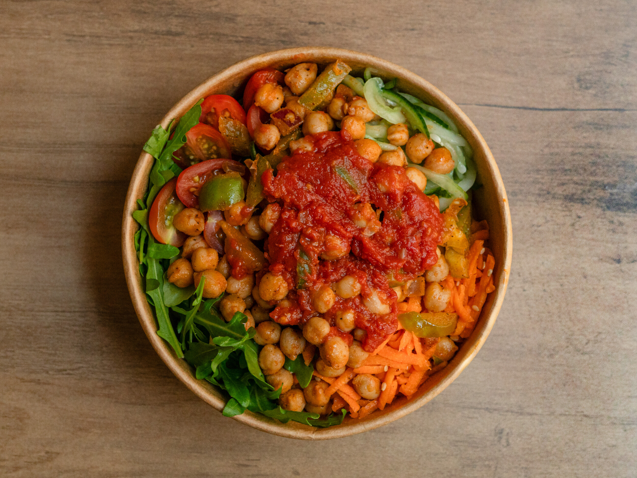 Draufsicht der veganen Ma Kanu Bowl von Mames Bowls & Wraps Gera: Eine nachhaltige Pappschale gefüllt mit Kichererbsen, einer würzigen Tomatensalsa, Karottenstreifen, Gurken, Rucola und Tomaten. Auch glutenfrei erhältlich.