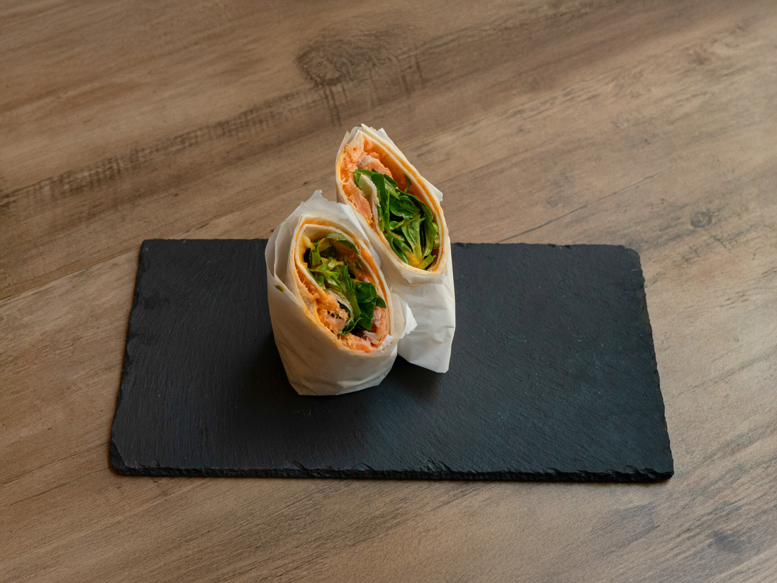 Der „Ma Owilli Ai Pai“ Lachs-Wrap von Mames Bowls & Wraps Gera, halbiert und in Pergamentpapier gewickelt, serviert auf einer schwarzen Schieferplatte. Im Inneren sind Räucherlachs, Feldsalat und Mango deutlich sichtbar. Auch glutenfrei erhältlich.
