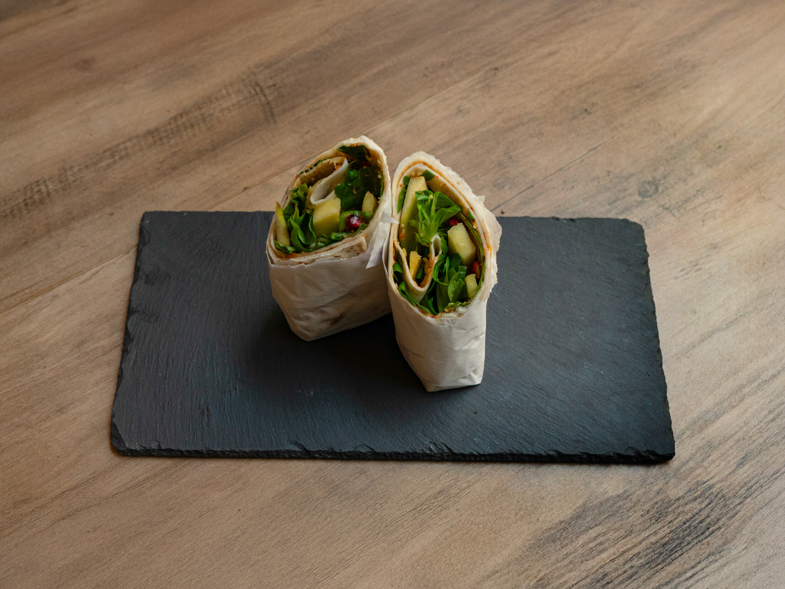 Der vegane „Ma Owilli Berena“ Wrap von Mames Bowls & Wraps Gera, halbiert auf einer Schieferplatte. Im Querschnitt sind Guacamole, Mango-Stücke, Feldsalat, Granatapfelkerne und Sesam gut zu erkennen. Auch glutenfrei erhältlich