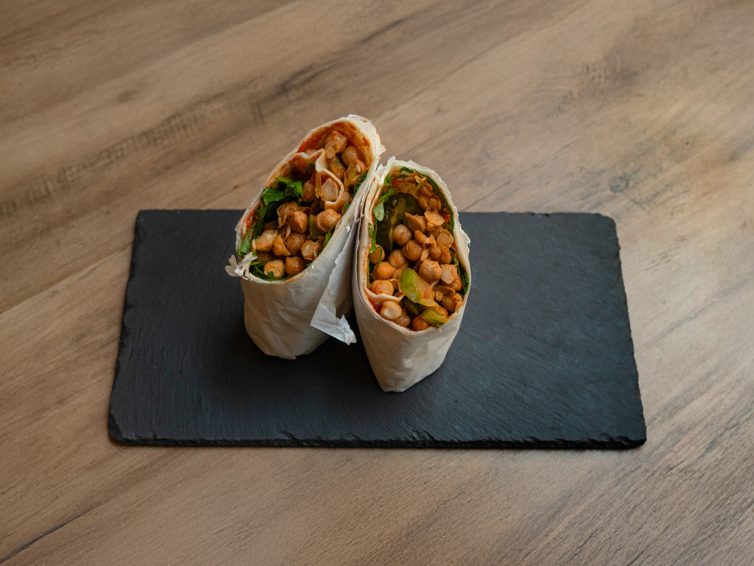 Draufsicht des halbierten „Ma Owilli Kanu“ Wraps von Mames Bowls & Wraps Gera: Der vegane Wrap liegt auf einer Schieferplatte und zeigt die Füllung aus Kichererbsensalat, Tomatensalsa und Rucola. Auch glutenfrei erhältlich.