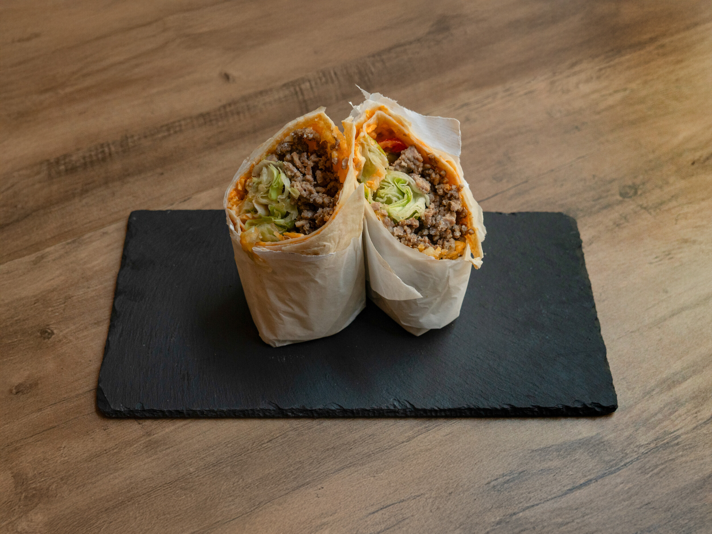 Der „Ma Owilli Kino“ Cheeseburger-Wrap von Mames Bowls & Wraps Gera, halbiert auf einer Schieferplatte. Der Wrap ist prall gefüllt mit saftigem Hackfleisch (Schwein/Rind), Eisbergsalat und geschmolzenem Käse. Auch glutenfrei erhältlich.