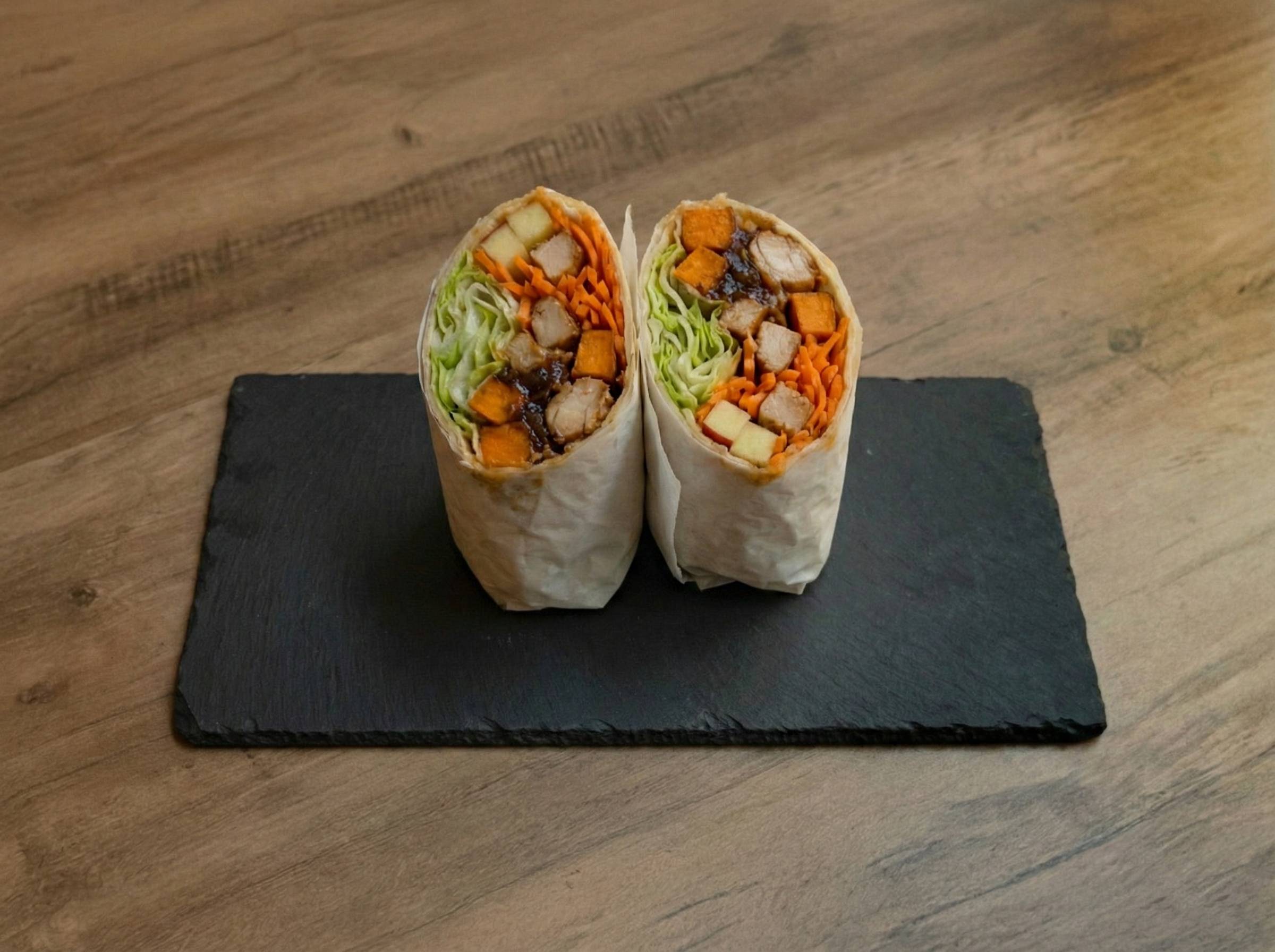 Nahaufnahme von zwei halbierten „Ma'Õwilli Inoa“ Wraps von Mames Bowls & Wraps Gera, angerichtet auf einer schwarzen Schieferplatte. Die Füllung zeigt goldene Süßkartoffeln, geraspelte Möhren, frischen Eisbergsalat, Apfelstückchen und saftiges Teriyaki-Hähnchen.